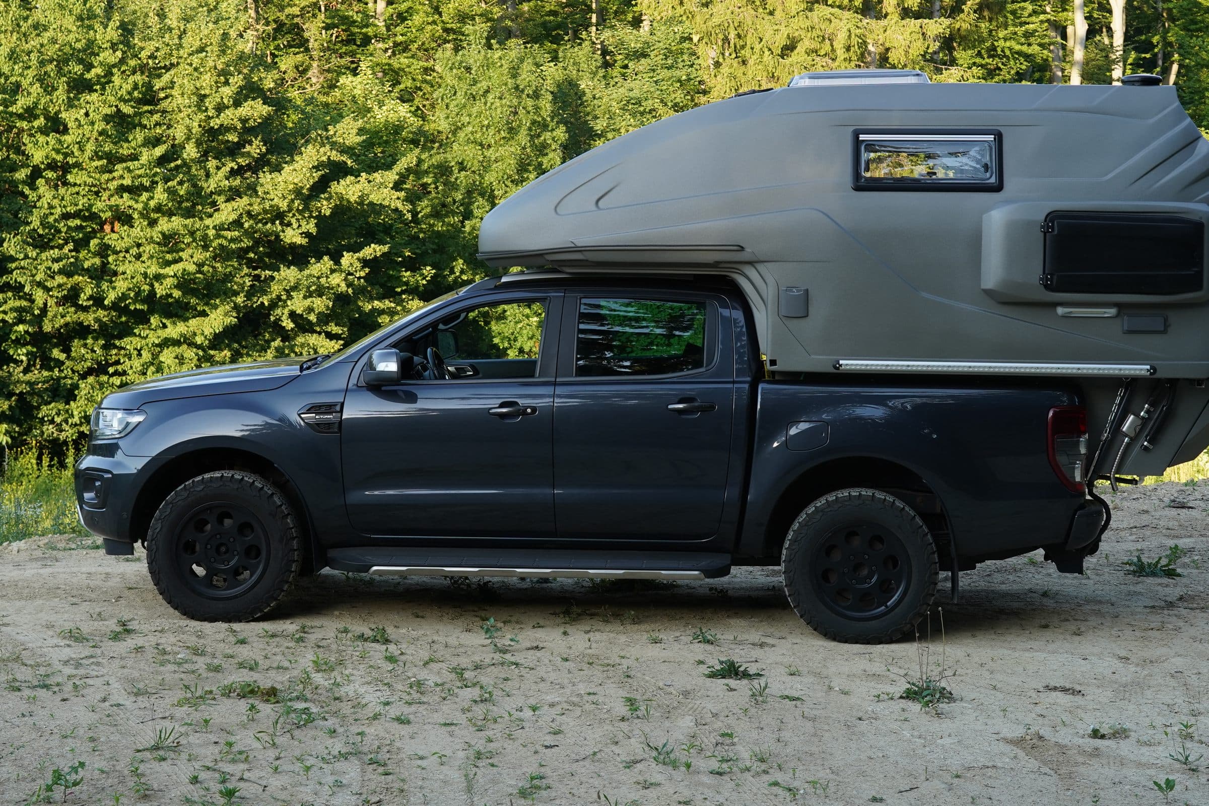 Olive-grey geocar camper on a dark pickup, side view in a sandy clearing with forest behind