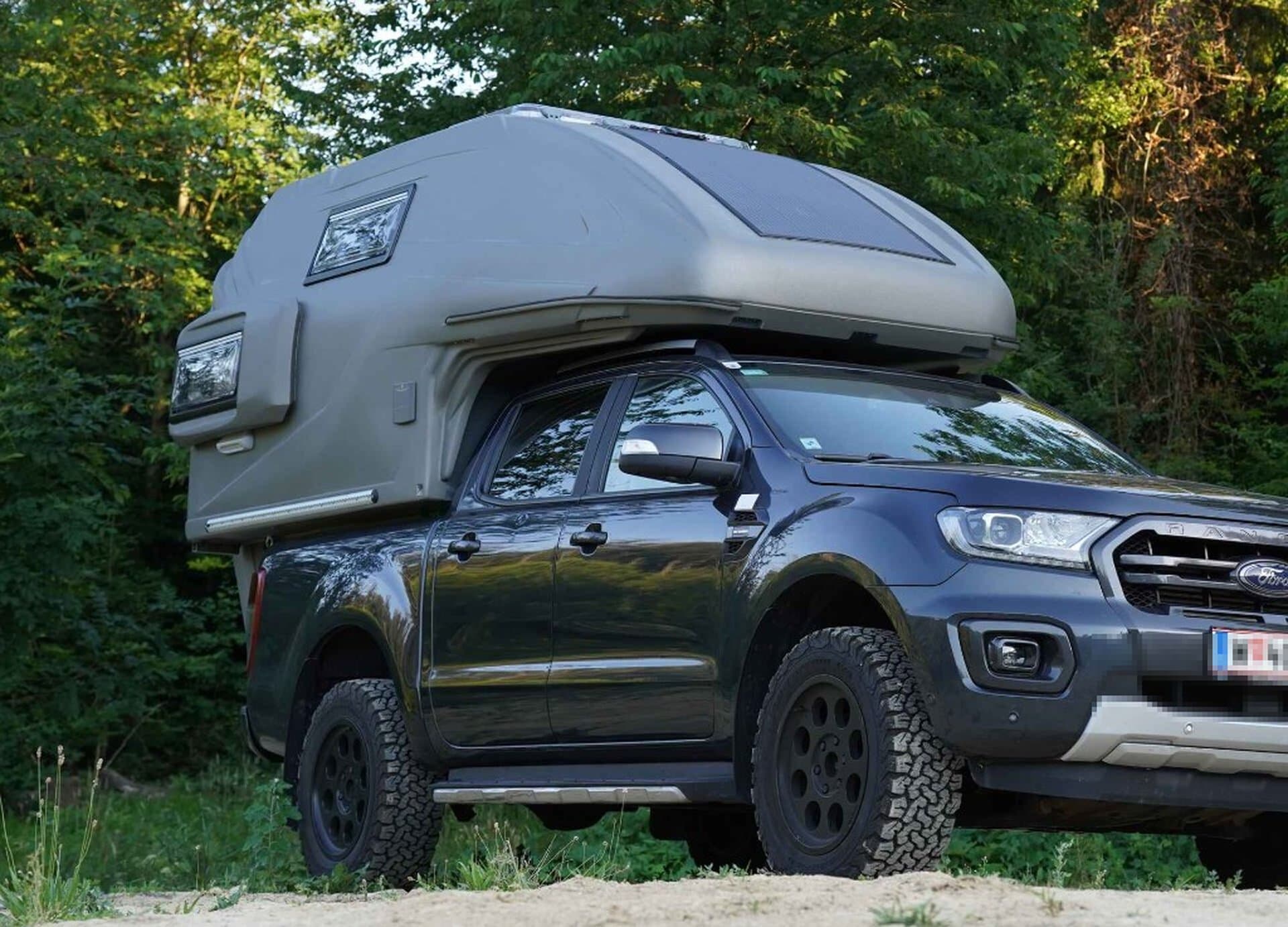 Dunkler Ford Ranger mit grauer geocar TAPIR Wohnkabine und Solarpanel vor Wald, Front-Schrägansicht