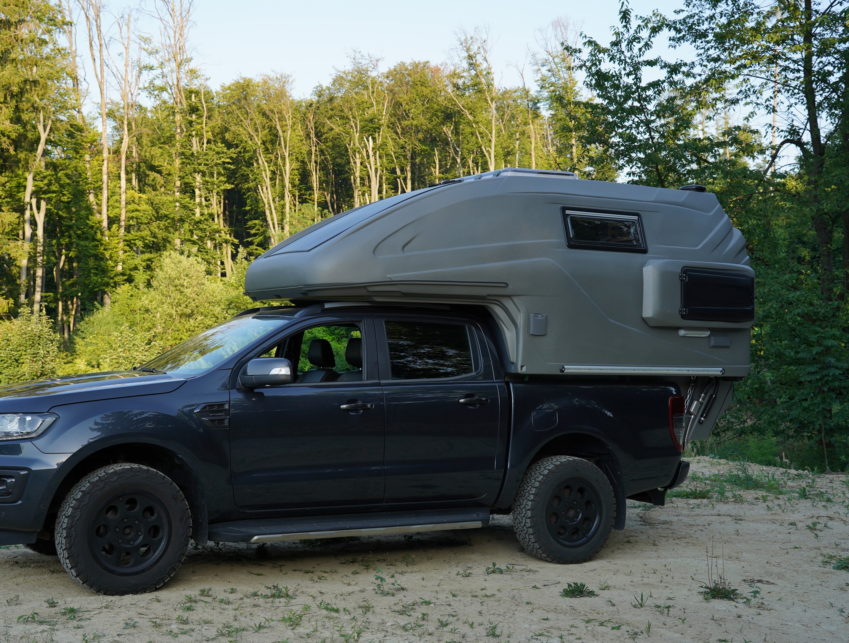 geocar TAPIR auf Ford Ranger Dunkler Ford Ranger mit grauer geocar TAPIR Wohnkabine und Solarpanel auf Sand vor Wald, Seitenansicht
