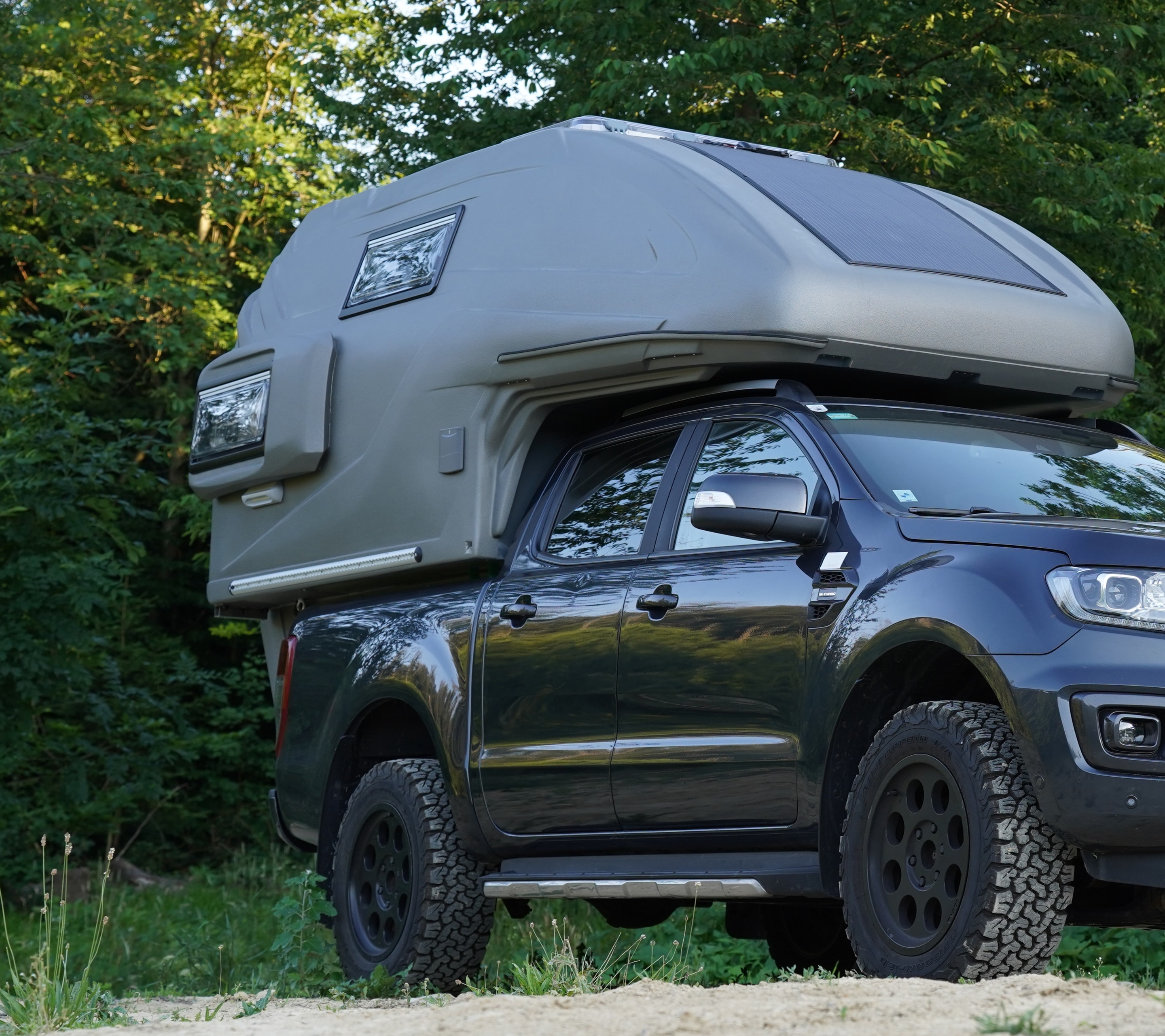 geocar TAPIR auf Ford Ranger Dunkler Ford Ranger mit grauer geocar TAPIR Wohnkabine und Solarpanel auf Sand vor Wald